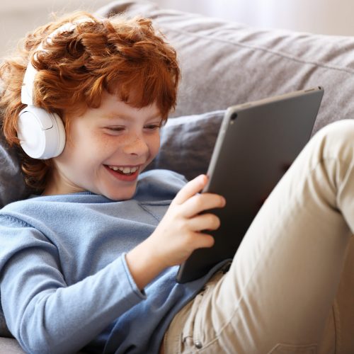 Cheerful little ginger boy in headphones smiling while sitting crossed legged on couch and using tablet at home